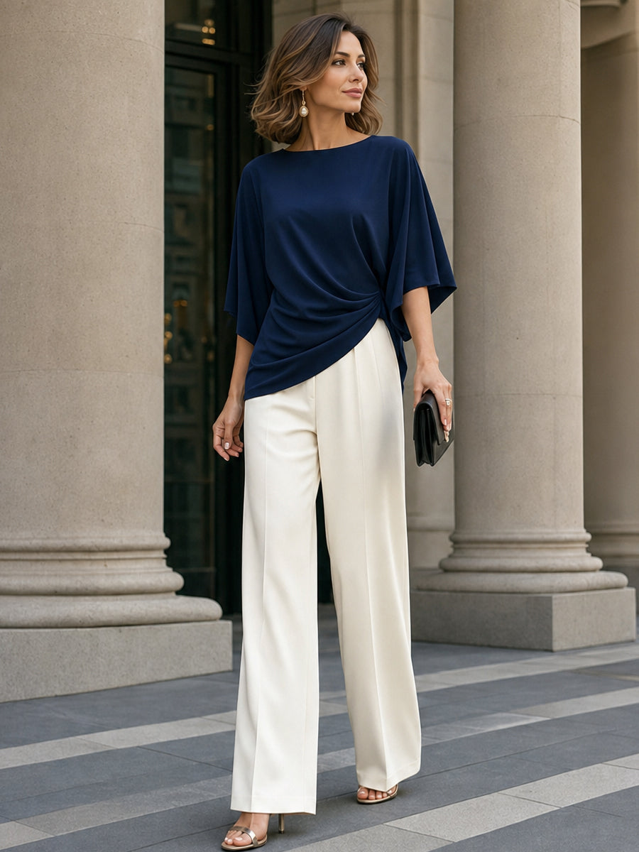 Model wearing the navy draped blouse with a pair of cream tailored wide-leg trousers, standing between stone columns. 13