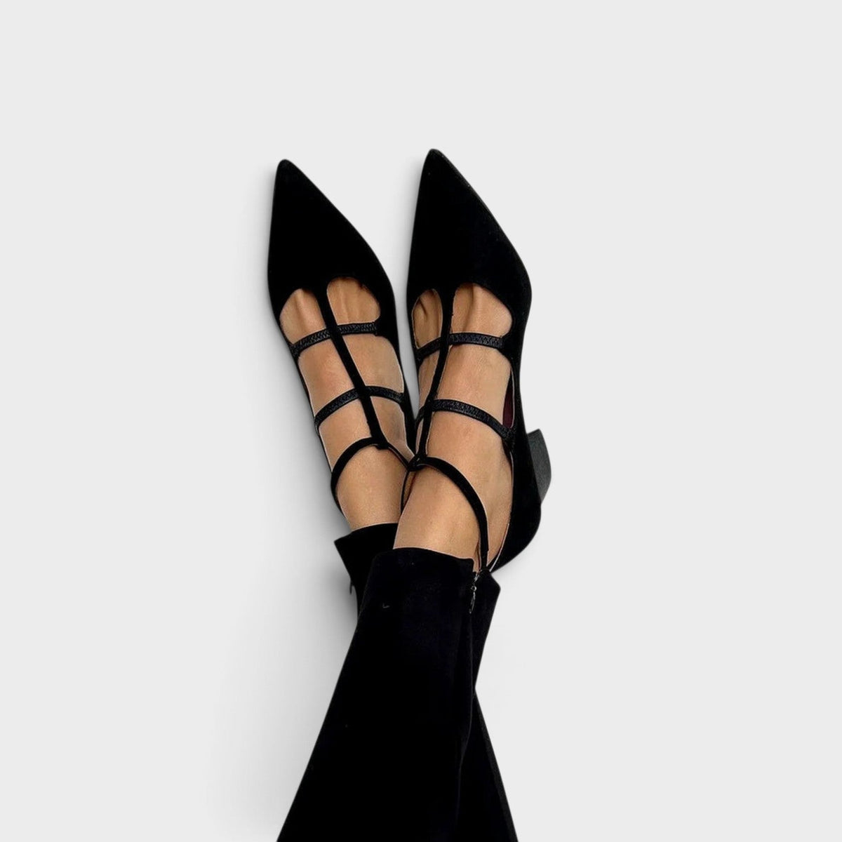Black pointed-toe heels with multiple thin cross straps and an ankle strap, worn with black trousers and feet crossed against a plain background. 1