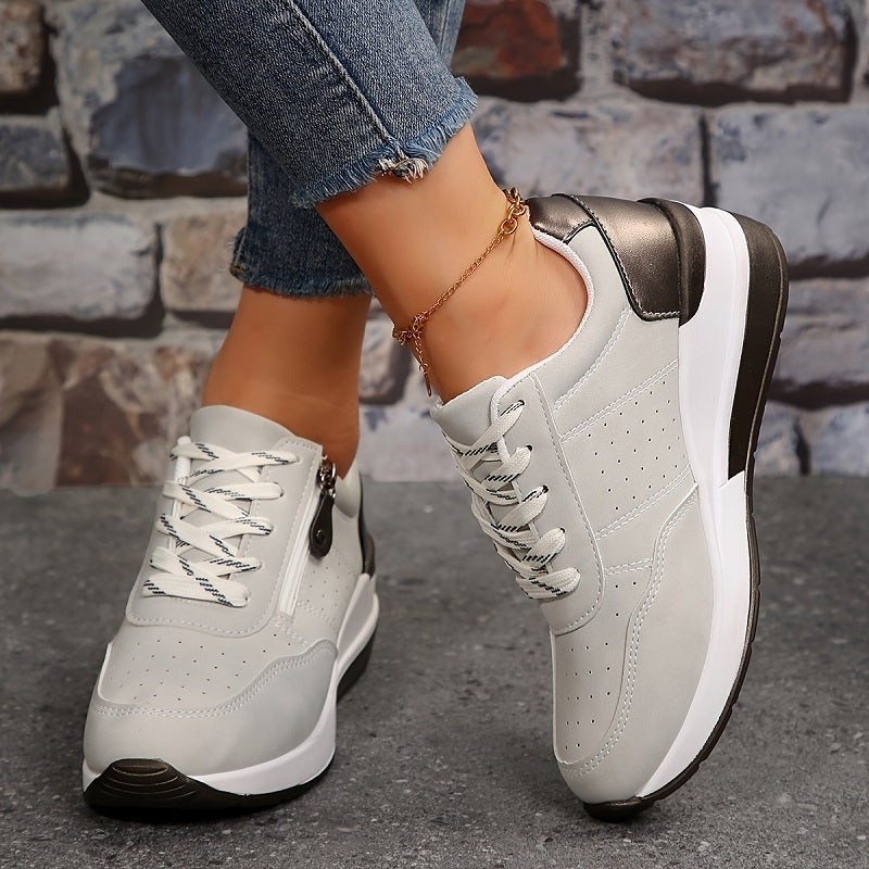 Light grey trainers worn on a brick backdrop, showing white laces, perforated panels and a thick sole 5