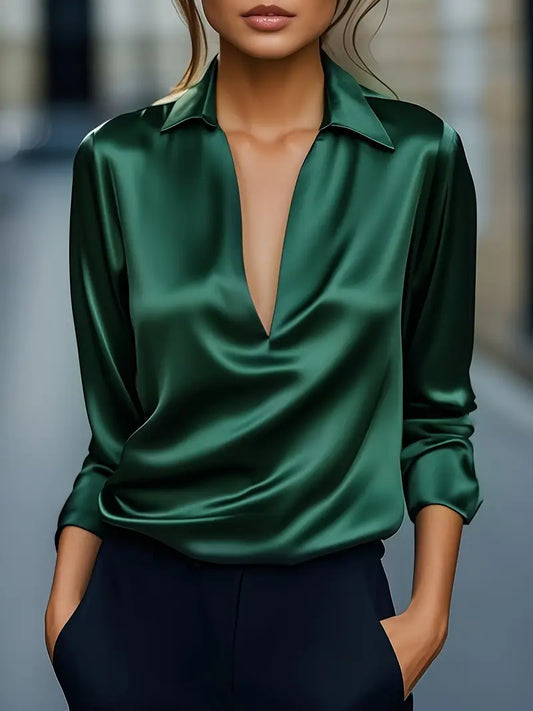 Model wearing the long-sleeved satin blouse in emerald green with navy trousers, hands in pockets, on a blurred street. 1