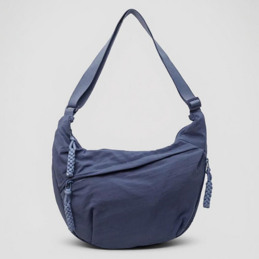 Front view of the blue crescent-shaped bag against a plain grey background, with a single strap, zip pockets and woven zip pulls. 1