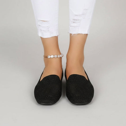 Front view of black sparkly knit slip-ons worn with white distressed jeans and a pearl anklet against a plain grey background. 2