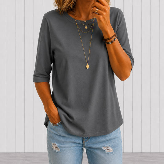 Woman wearing the round-neck half-sleeve t-shirt in dark grey, layered gold necklaces and ripped light-wash jeans, against a pale panelled wall. 1