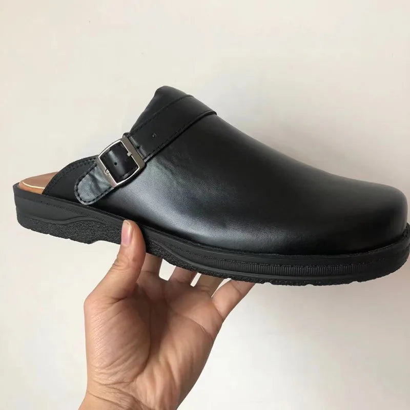 Black clog with a smooth finish, silver buckle strap and black sole, held in one hand against a plain wall. 5