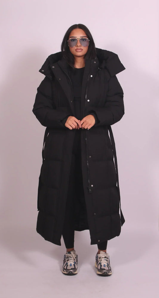 Front view of the black quilted coat worn open with the belt hanging loose, model's hands at the waist 2