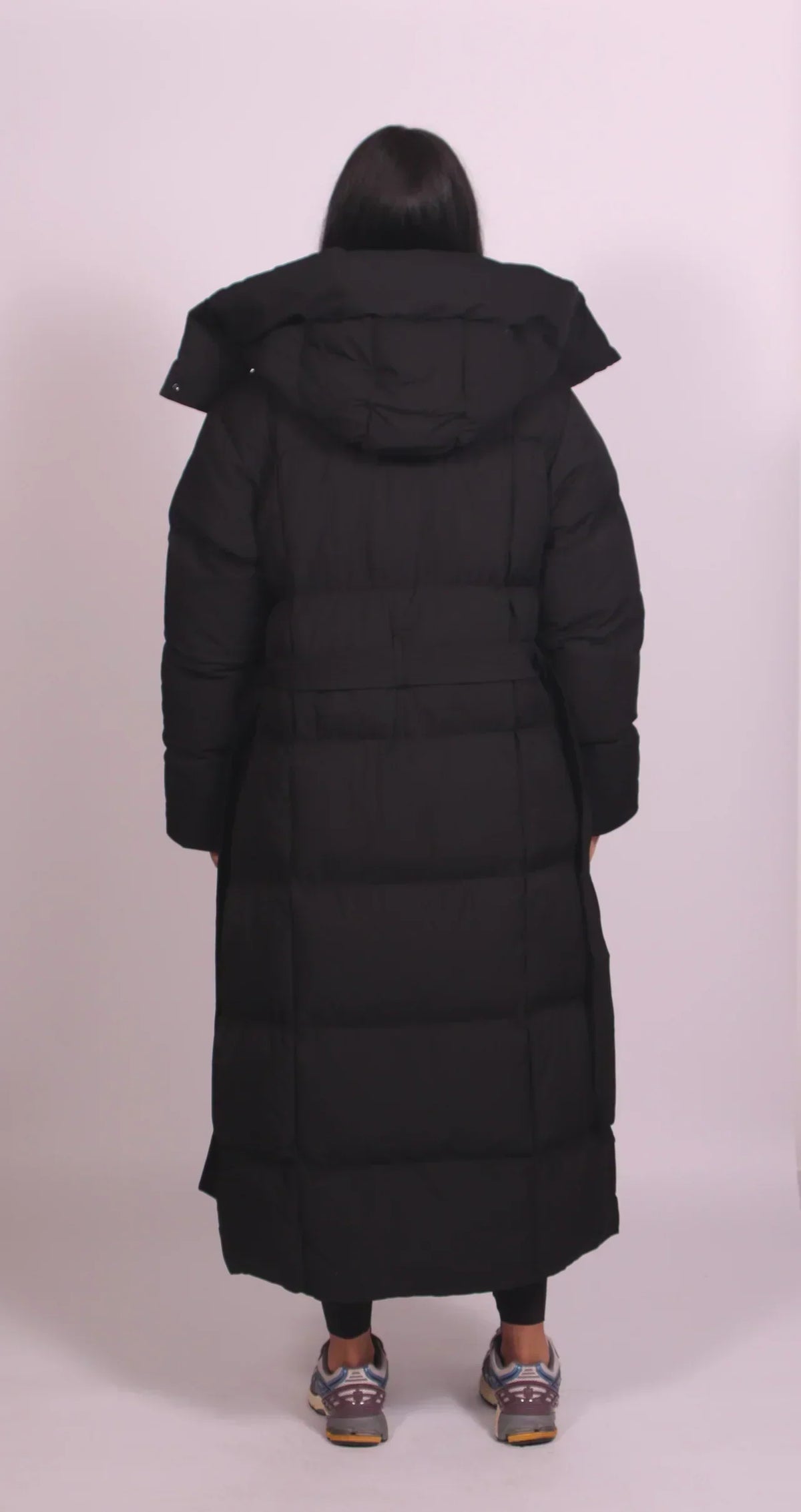 Back view of the black padded coat, showing the hood, horizontal quilting and waist belt on a model 4