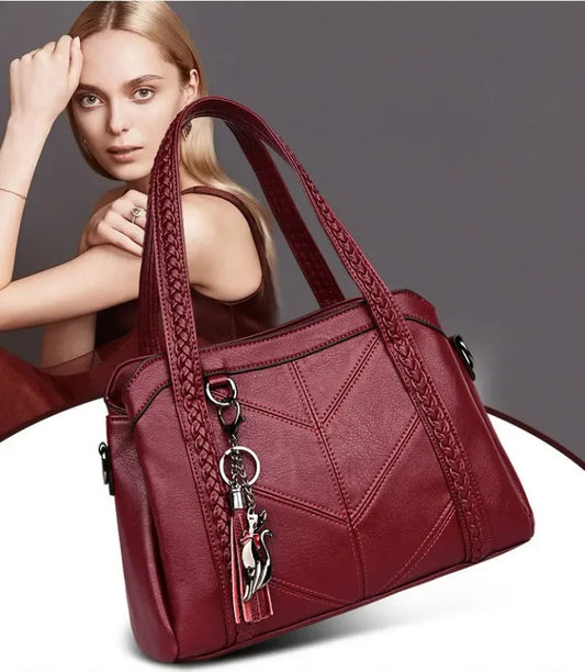 Burgundy tote with braided handles and cat charm, shown beside a blonde model posing with her hand raised to her head. 1