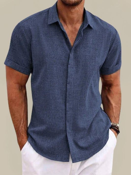 Model wearing the textured short-sleeved shirt in navy blue, buttoned with a collar, hands in pockets of white trousers. 1