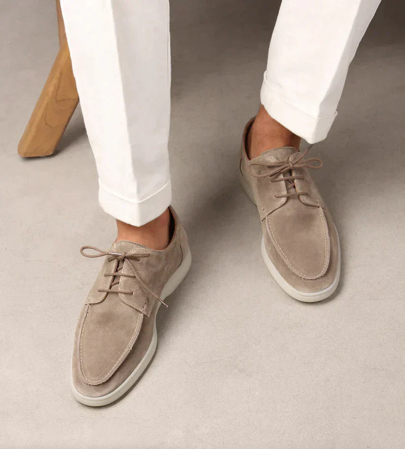 Man's legs in white cuffed trousers wearing taupe suede lace-up loafers with white soles, viewed from above on a grey floor. 2