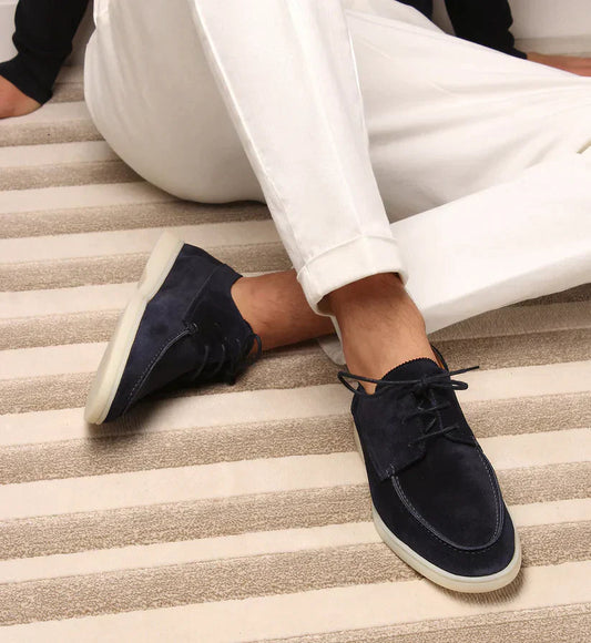 Man seated on a striped rug wearing navy suede lace-up loafers with pale soles, styled with white cuffed trousers. 1