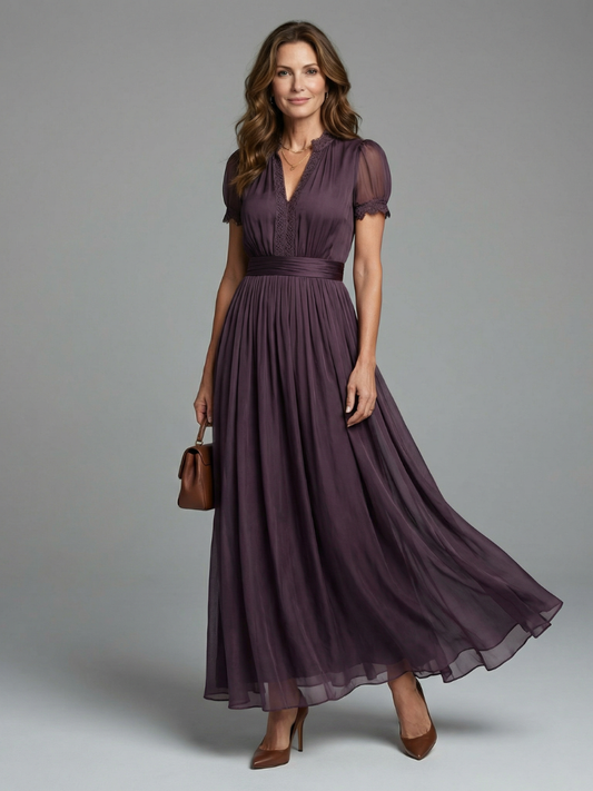 Front view of a woman in a plum purple maxi dress with lace-trimmed V-neck, sheer puff sleeves and pleated skirt, holding a brown handbag. 1