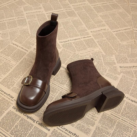 Brown suede ankle boots with glossy leather toe caps and brass buckles, one boot lying on its side to show the ridged rubber sole. 1