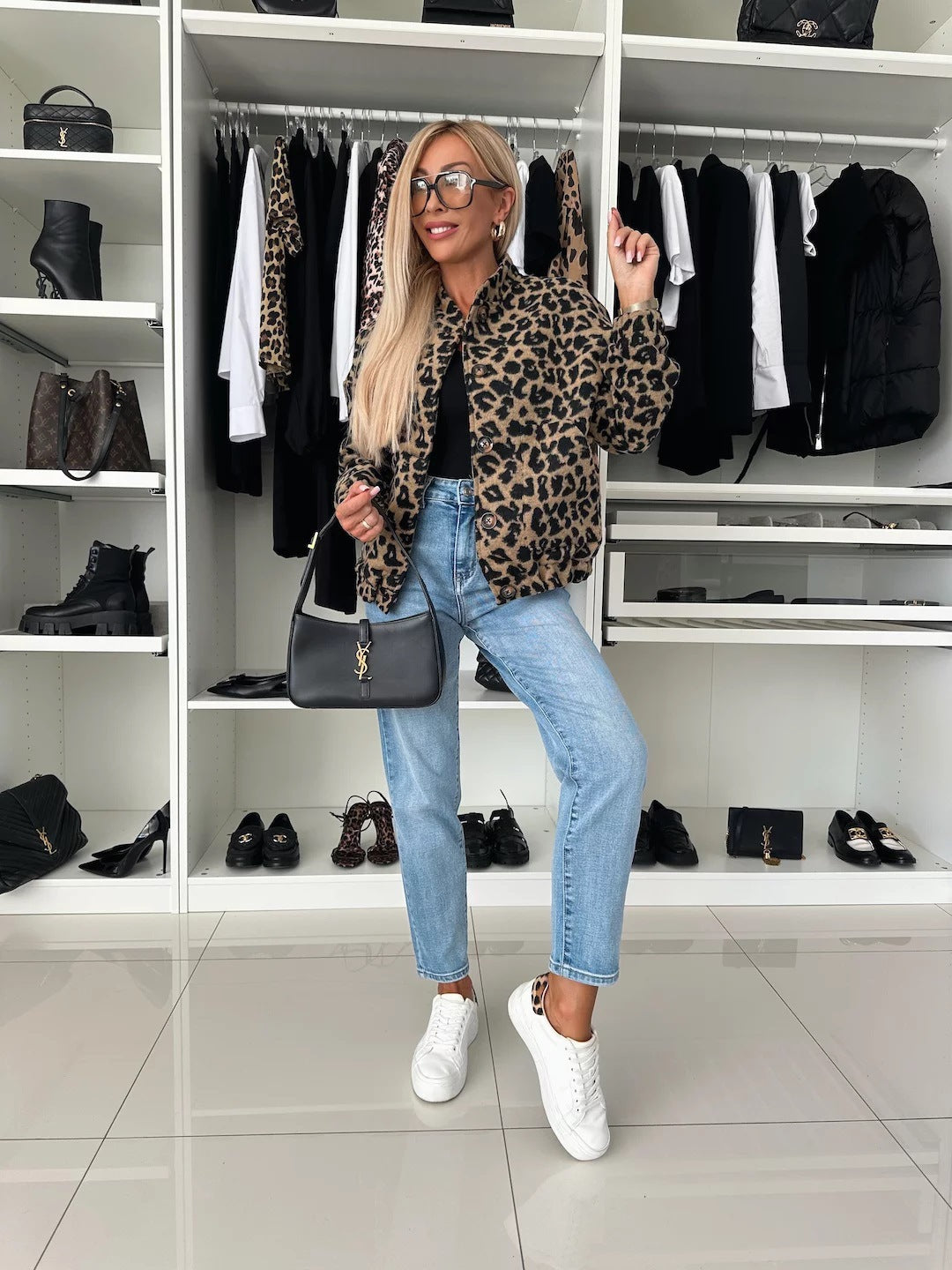 Full-length view of a model in the leopard print bomber jacket, black top, light jeans and white trainers, holding a black shoulder bag. 5