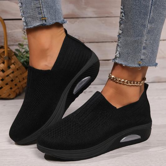Black knit slip-on shoes with a thick black sole and visible air cushion, worn with frayed jeans and a gold ankle chain. 1