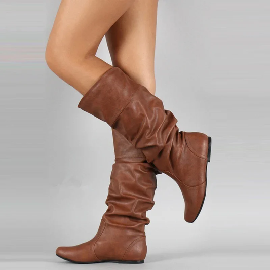 Model's raised leg in tan-brown slouchy flat boots with a folded-over cuff, shown in side profile against a grey backdrop. 1