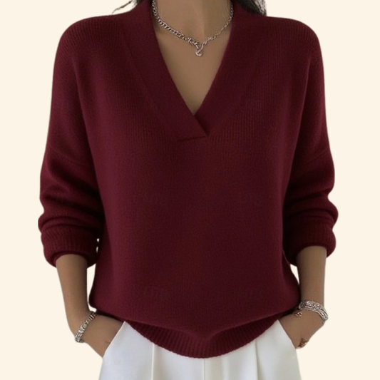 Model wearing the knitted V-neck jumper in burgundy with white trousers, silver necklace and bracelets, hands in pockets. 1