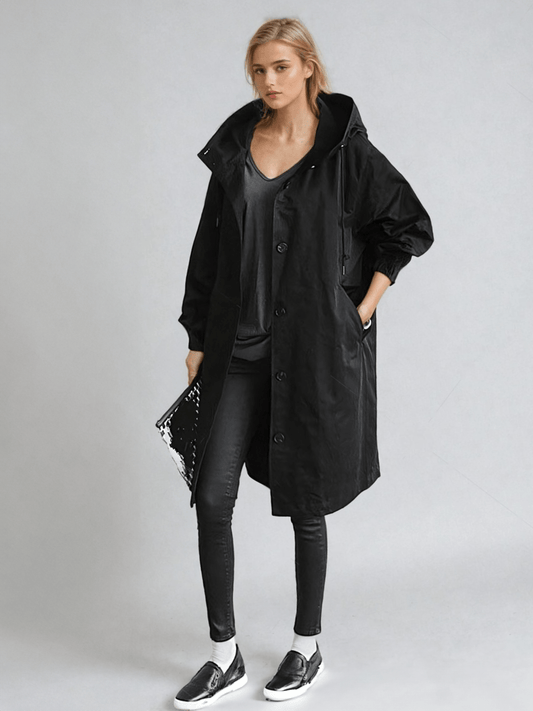 Model in the black hooded coat with skinny trousers and slip-on trainers, holding a clutch 2