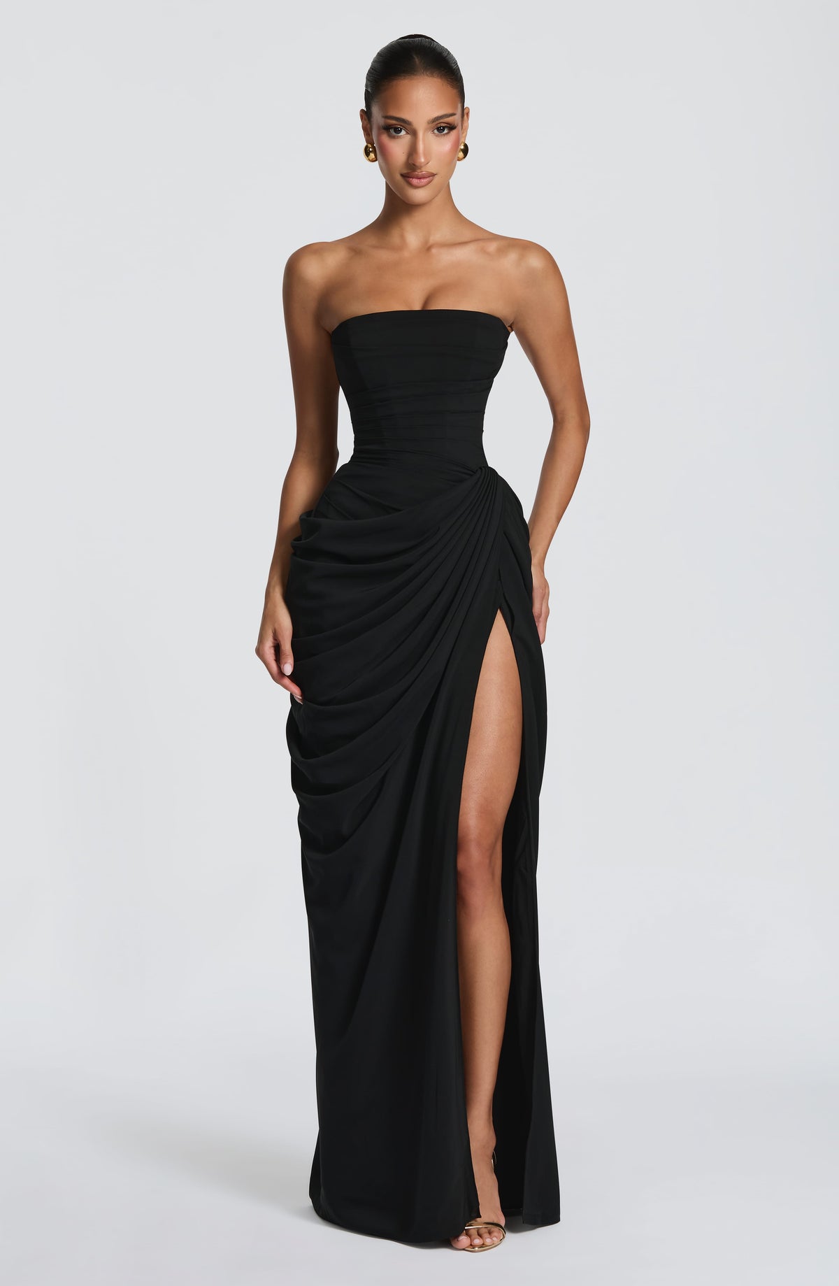 Front view of the black dress with one hand on the draped hip, high slit open and gold heels. 34