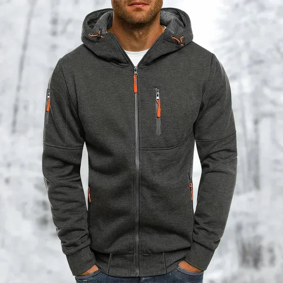 Model wearing the hoodie in dark charcoal grey with orange zip pulls, ribbed cuffs and hem, against a snowy backdrop. 9