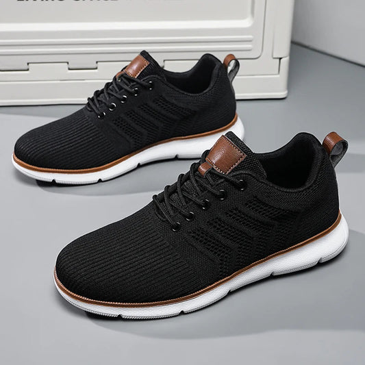 Pair of black knitted mesh trainers with white soles and tan leather trim, shown on a grey floor beside a storage box. 1