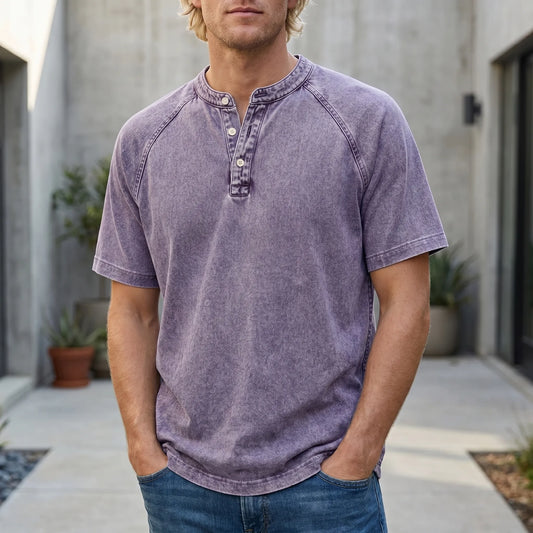 Fair-haired man in a faded purple raglan-sleeve Henley with hands in jeans pockets, standing outdoors in a concrete courtyard. 1