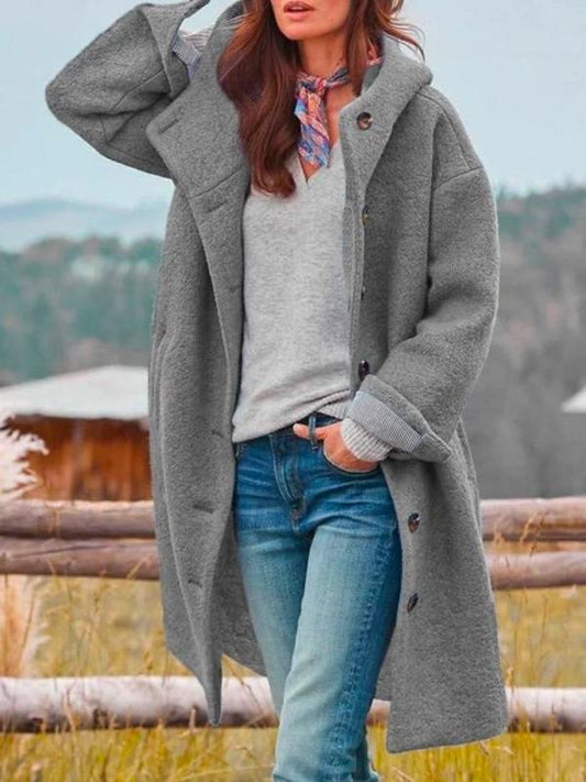 Woman in the knee-length hooded coat in grey, worn open over a grey jumper and jeans with a patterned neck scarf. 2