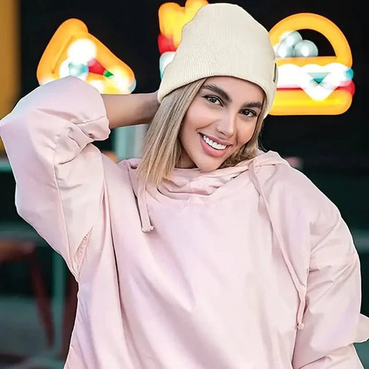 Smiling woman in a cream knit beanie and pale pink hooded top, hand behind head 2