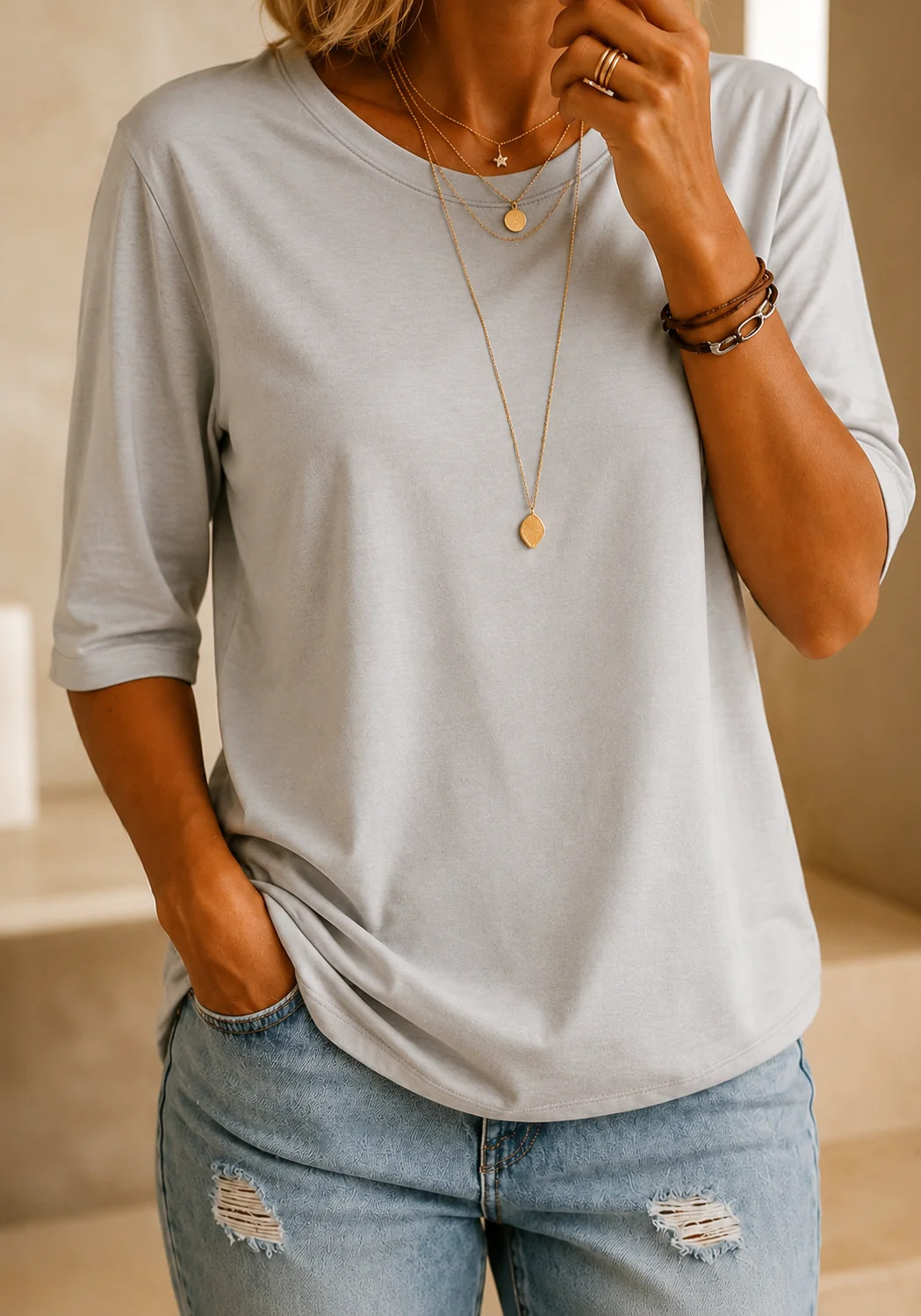 Model wearing the tee in light grey with layered gold necklaces and ripped jeans, in a warm neutral interior. 13
