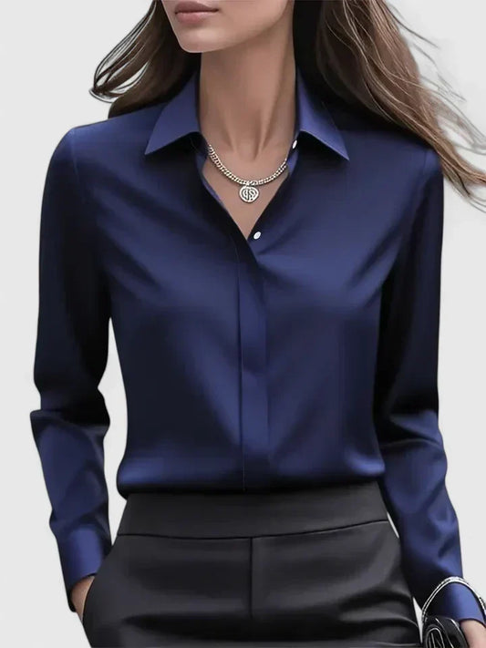 Front view of the navy satin blouse on a model, with pointed collar, concealed placket and a silver necklace, worn with black trousers. 1