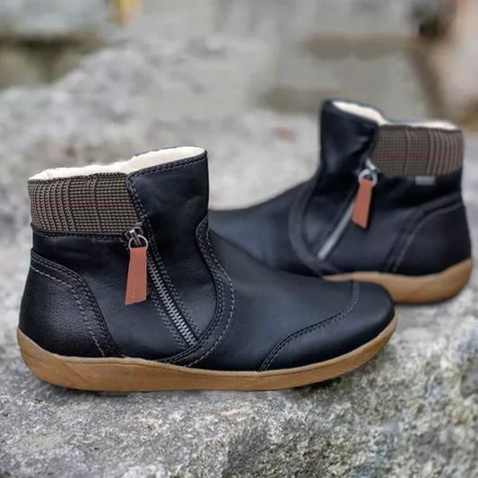 Black version of the ankle boots with side zip and orange-brown pull tab, houndstooth collar and tan sole, on a stone surface. 2
