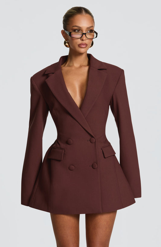 Chocolate-brown double-breasted blazer dress worn by a model in clear-lens glasses, front view with deep plunge neckline and fabric-covered buttons. 1