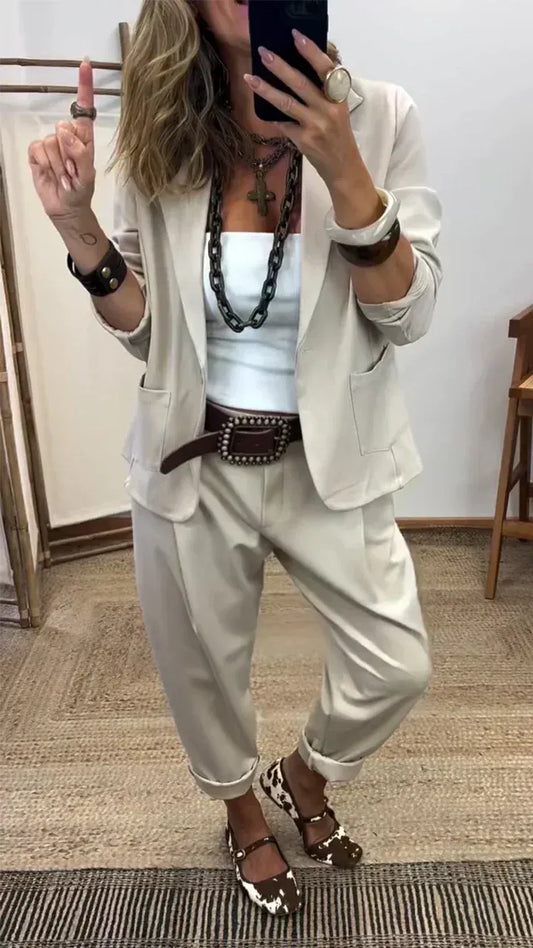 Woman wearing the light stone-coloured blazer with patch pockets and matching rolled-hem trousers, styled with a chunky chain necklace and wide belt. 2