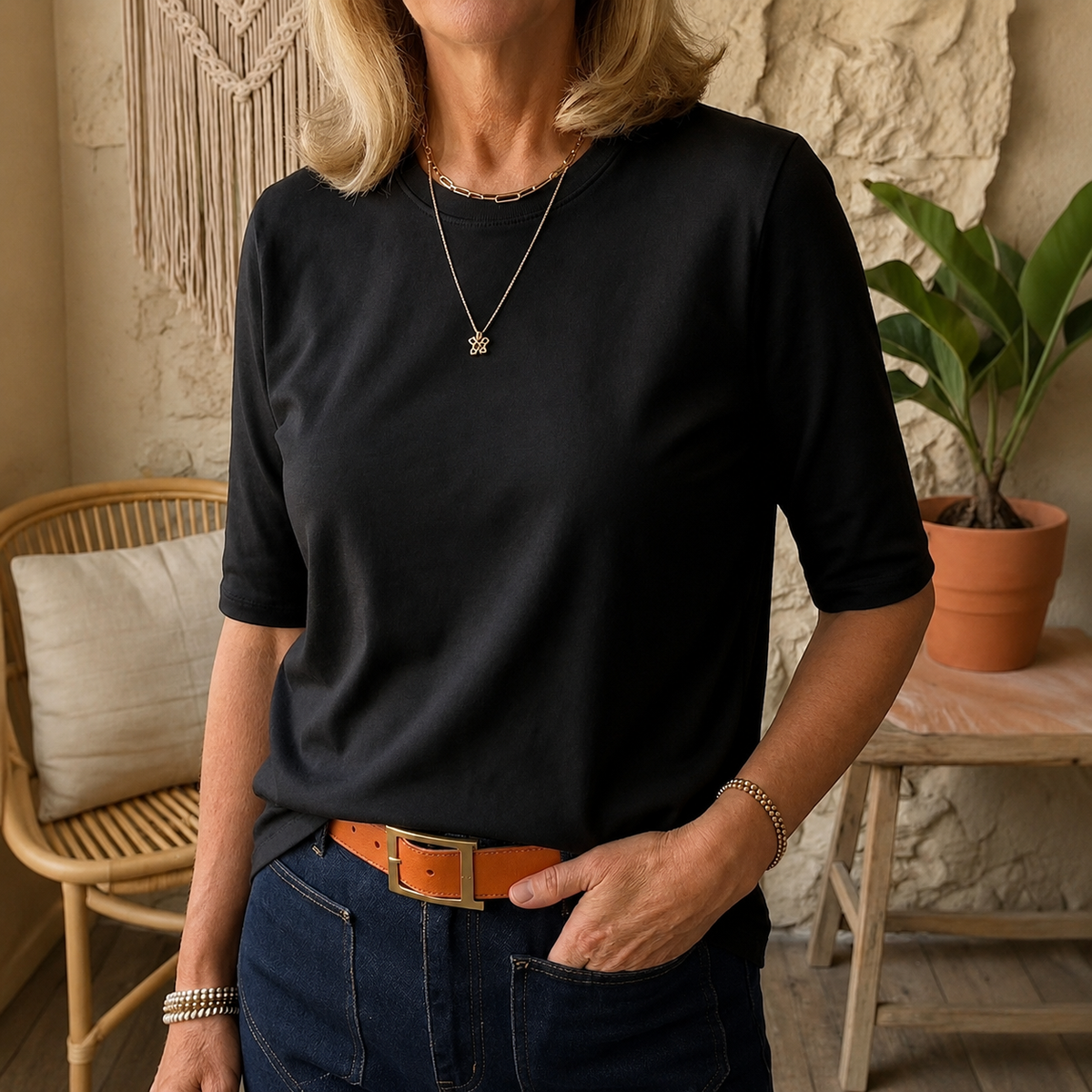 Woman wearing the round-neck half-sleeve t-shirt in black, tucked into dark jeans with a tan belt, in a cosy room with a rattan chair. 7