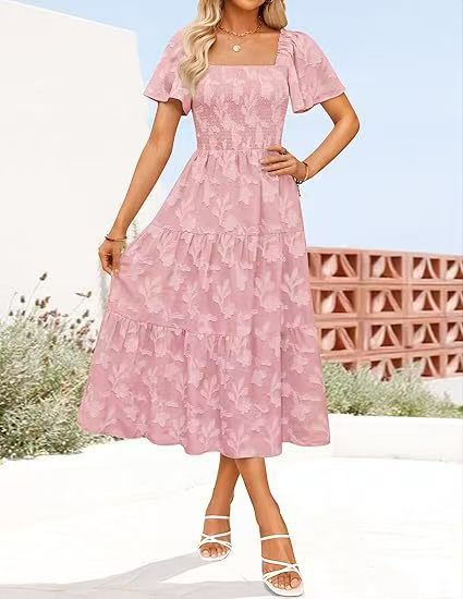 Model wearing the dress in soft pink, smocked bodice and flutter sleeves, outdoors by a terracotta wall 2
