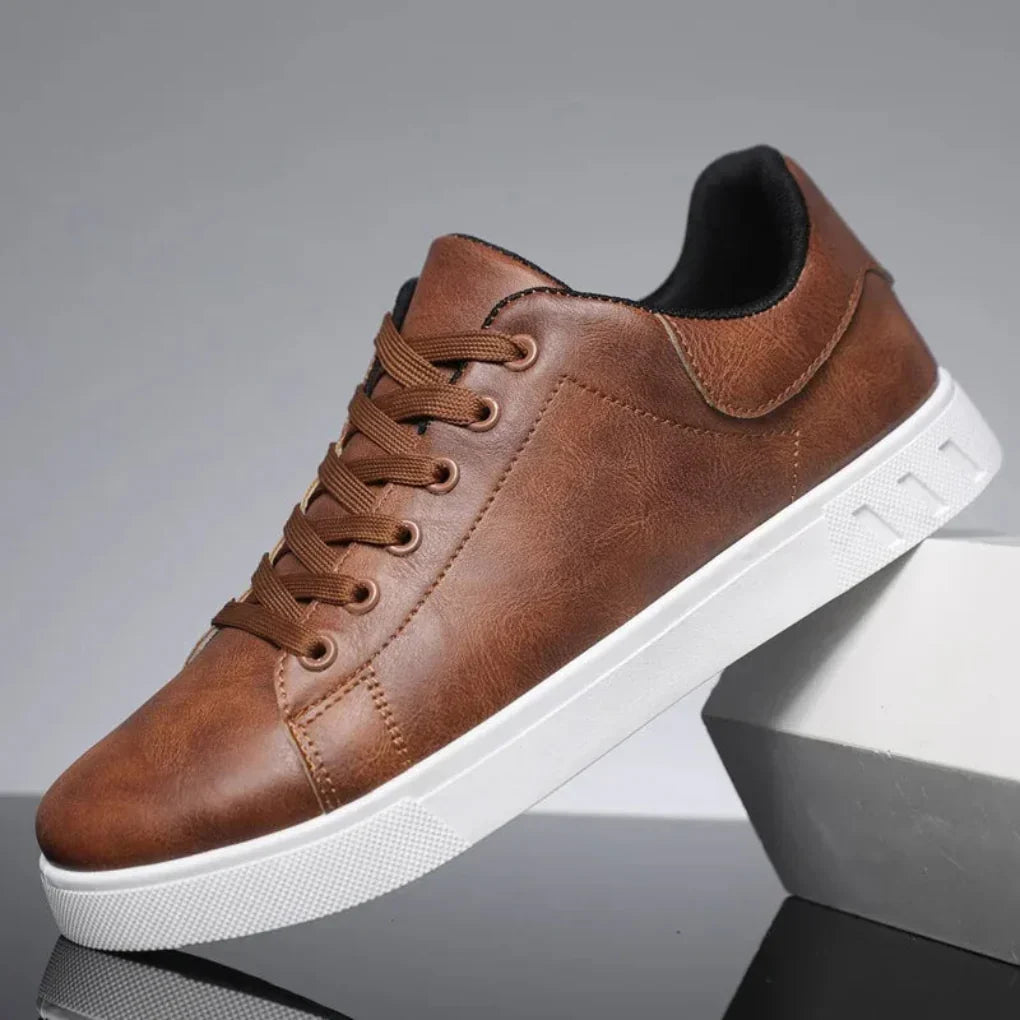 Side view of a low-top lace-up shoe in tan-brown faux leather with a thick white sole, resting on a grey block. 1