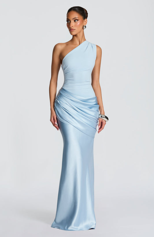 Model in a pale blue one-shoulder maxi dress with a draped satin hip panel, head turned to the side. 1