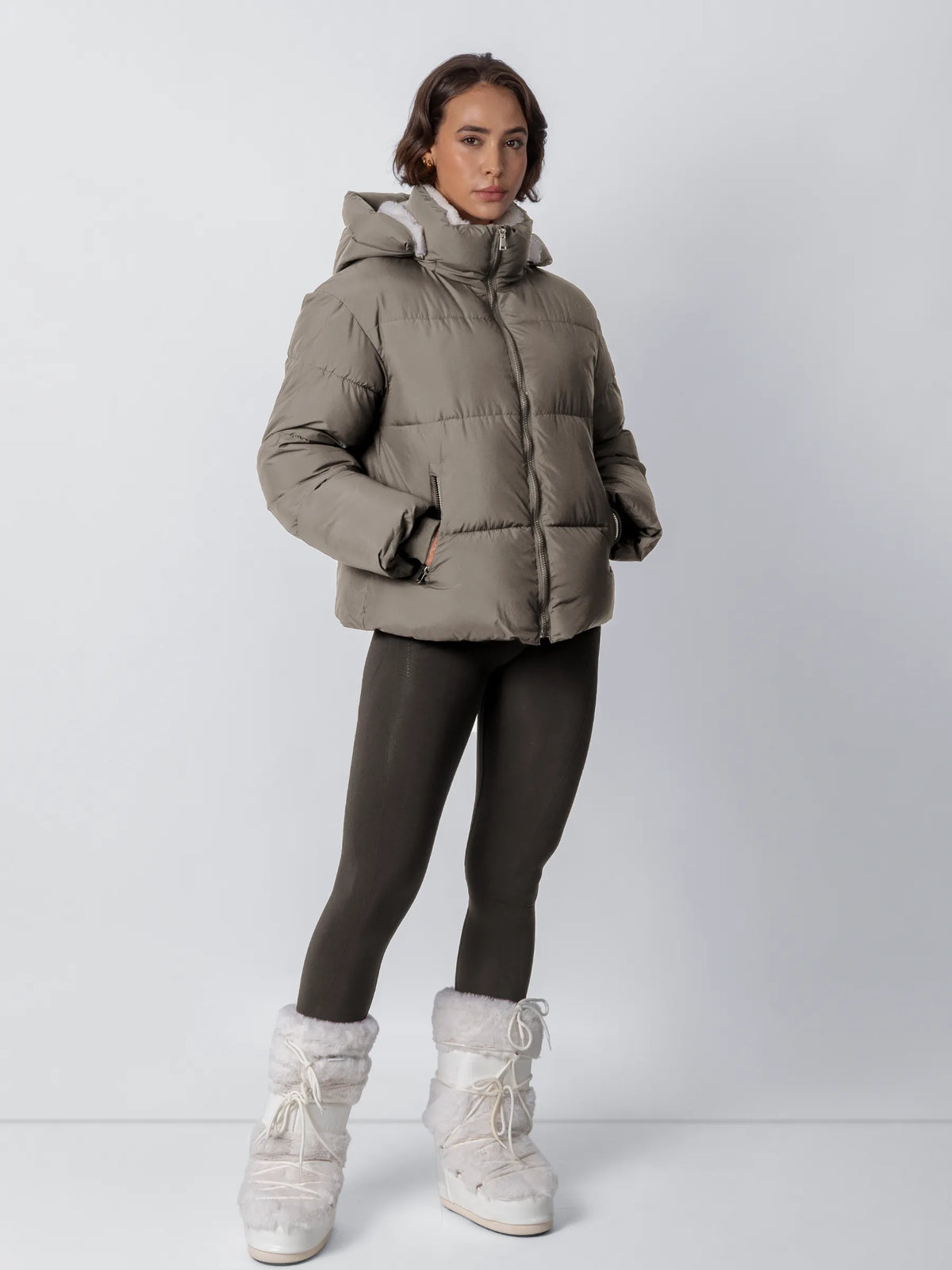 Full-length view of a model in a taupe puffer jacket with fleece collar, dark leggings and white fur boots 21