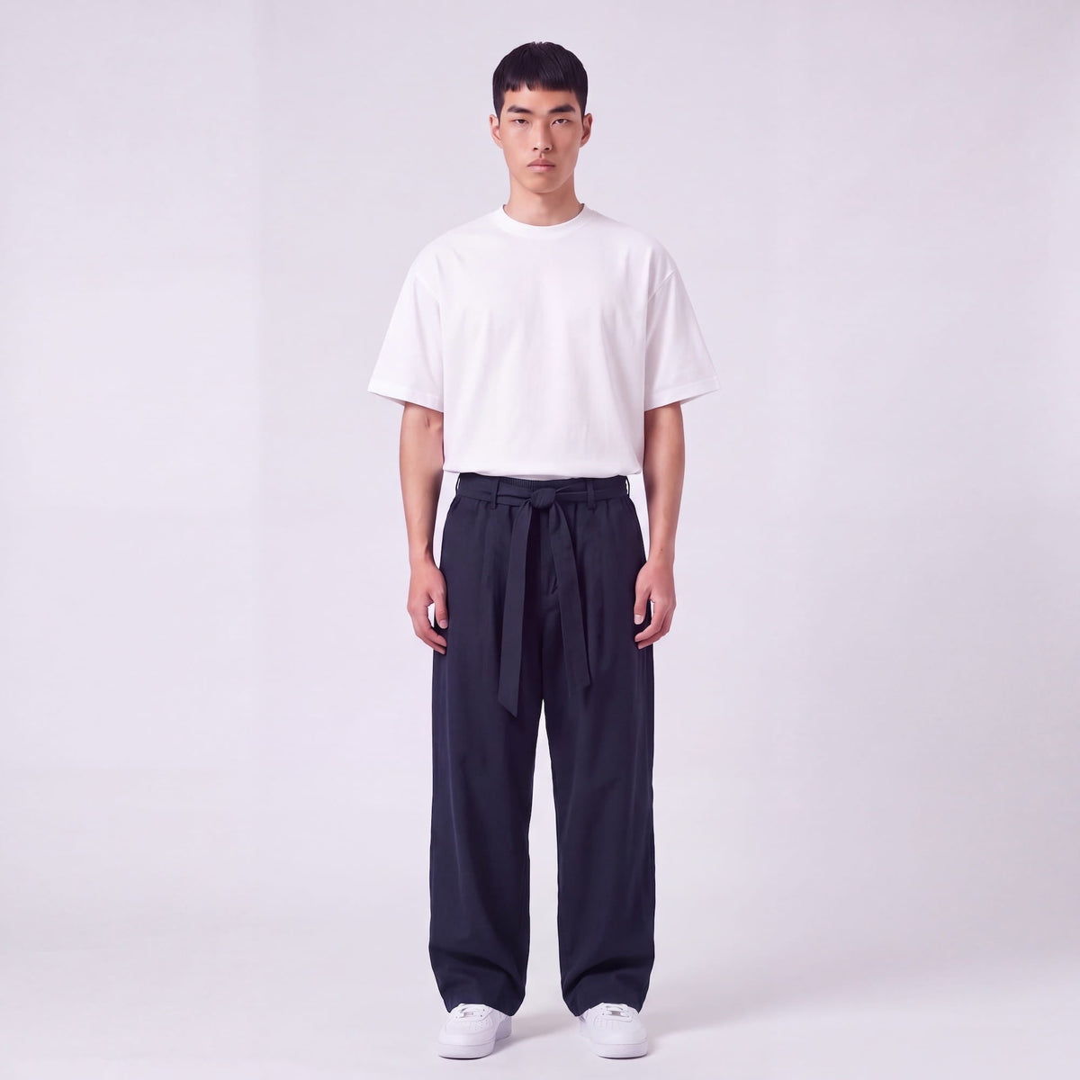Full-length front view of a model wearing navy trousers with the waist belt tied in a knot, with a white tee. 2