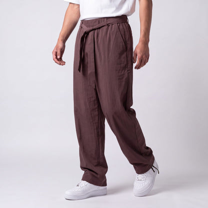 Angled three-quarter view of plum-brown trousers with tied belt and drawstring ankle, showing the loose, draped leg while walking. 8
