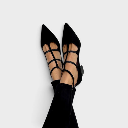 Black pointed-toe heels with multiple thin cross straps and an ankle strap, worn with black trousers and feet crossed against a plain background. 1