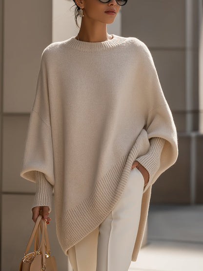 Beige knit poncho worn by a model against stone pillars, showing the wide batwing sleeves and ribbed hem 2
