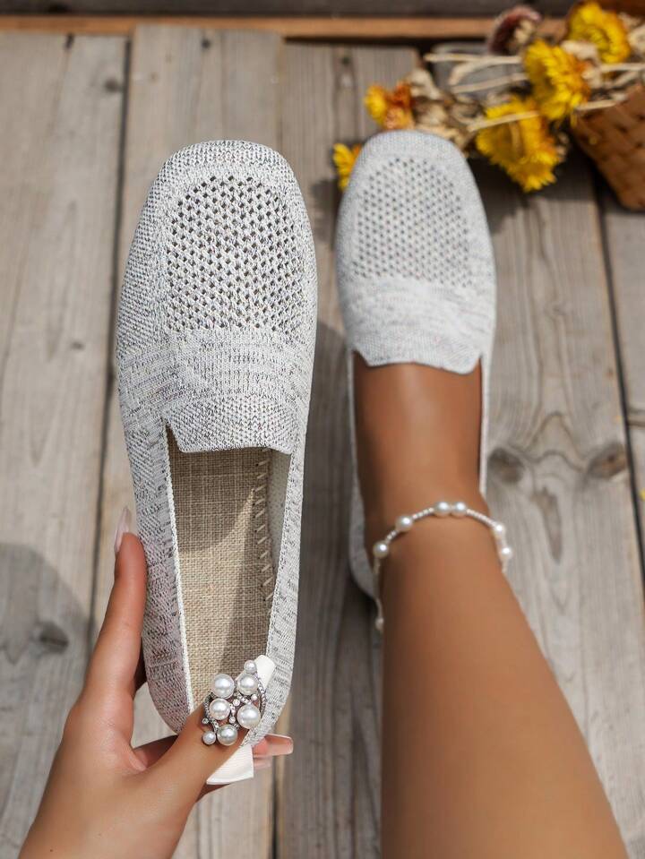 Top view of light grey perforated knit slip-ons on wooden boards, one held to show the woven footbed and heel tab. 15