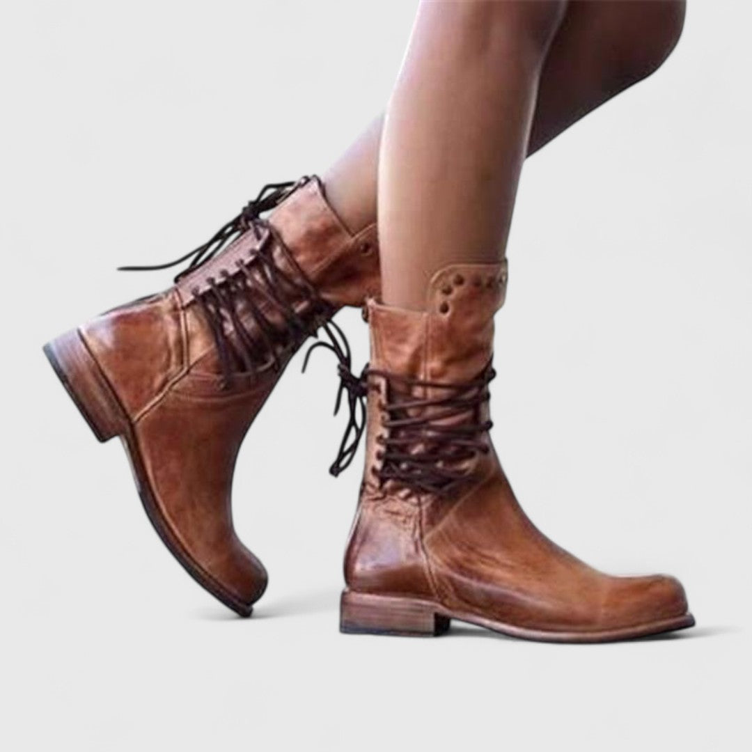 Model wearing worn-look brown leather boots with dark criss-cross lacing, walking with one foot raised against a pale background. 8