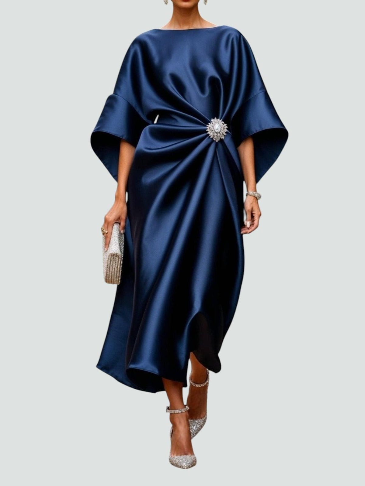 Model wearing the satin dress in deep navy, with wide cape sleeves, gathered waist, crystal brooch and glittering clutch and heels. 1
