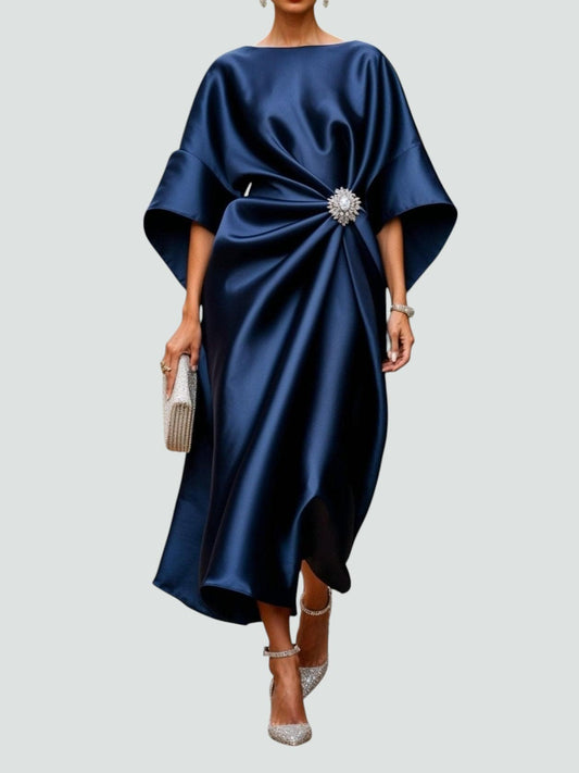 Model wearing the satin dress in deep navy, with wide cape sleeves, gathered waist, crystal brooch and glittering clutch and heels. 1