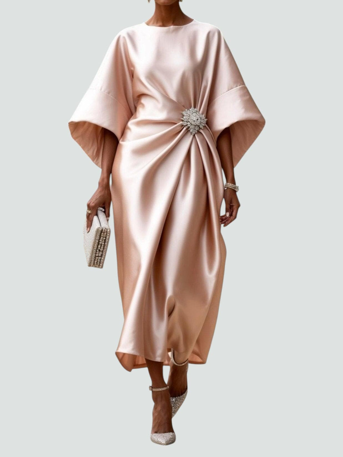 Model wearing the satin dress in soft blush pink, with wide sleeves, ruched side drape and a crystal brooch, holding a beaded clutch. 5