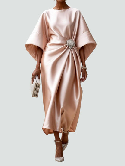 Model wearing the satin dress in soft blush pink, with wide sleeves, ruched side drape and a crystal brooch, holding a beaded clutch. 5