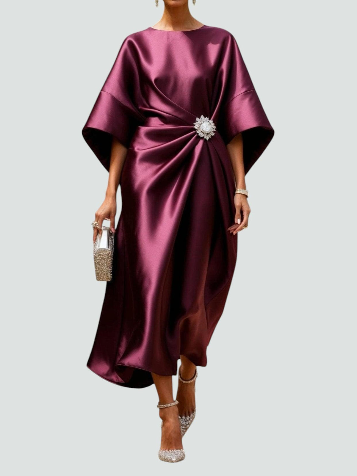 Model wearing the glossy satin dress in burgundy, with flared sleeves, draped waist and a sparkling brooch, holding a jewelled clutch. 2