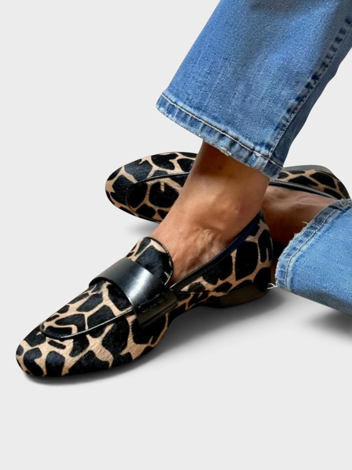 Side view of the animal-print loafers worn with blue jeans, one foot crossed over the other, showing the black leather strap. 1
