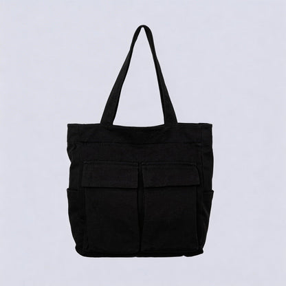 Black canvas tote bag seen from the front, with two large flap pockets, side pockets and two upright handles, against a plain pale background. 1